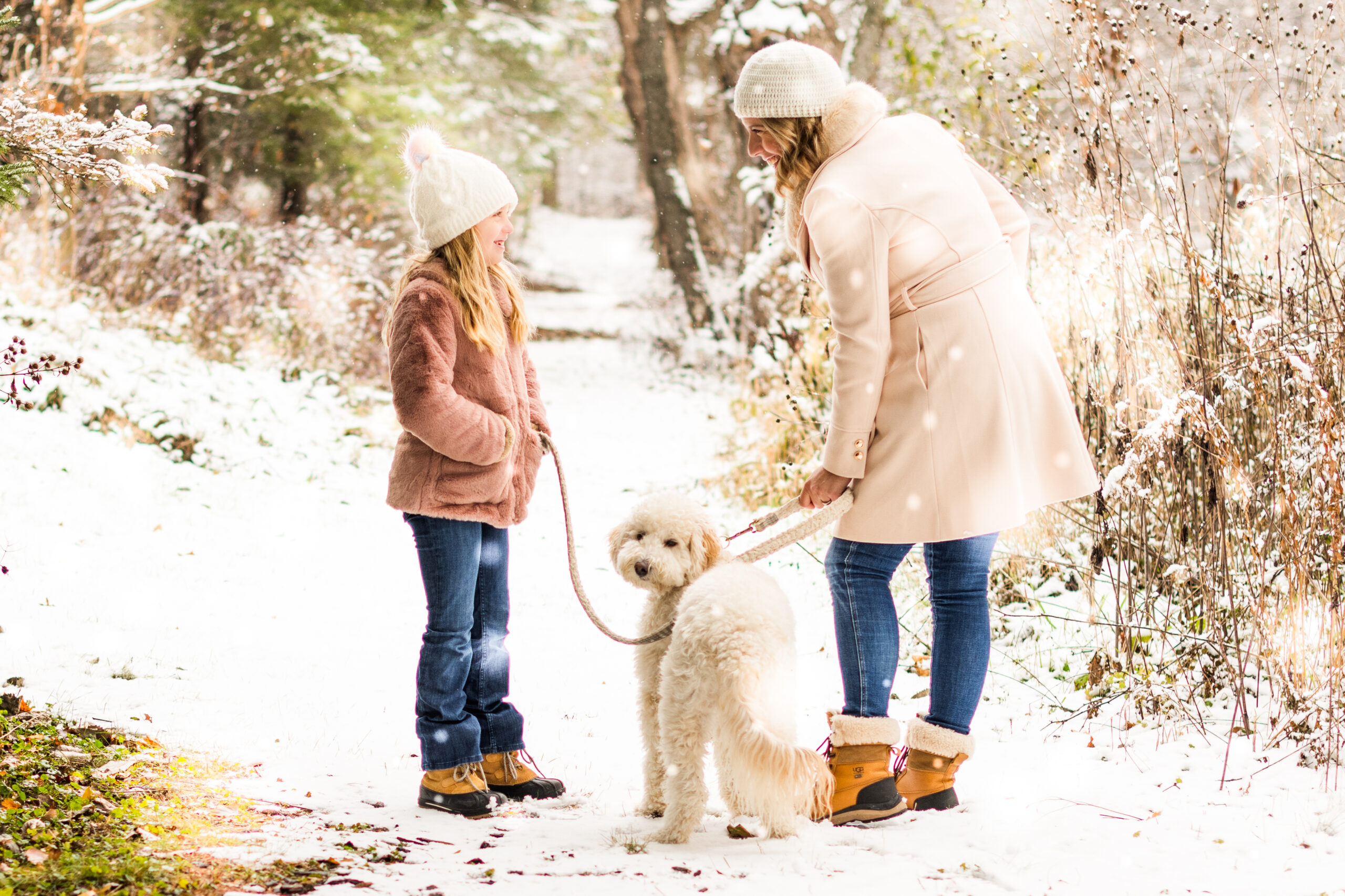 Mom, daughter, and fluffy white dog share a joyful snowy moment. Smiles, laughter, and warmth in the winter wonderland with evergreen trees in the background.