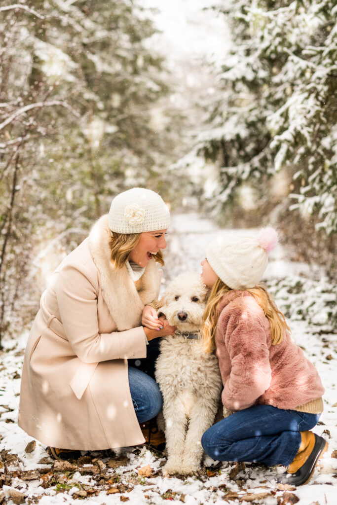 Mom, daughter, and fluffy white dog share a joyful snowy moment. Smiles, laughter, and warmth in the winter wonderland with evergreen trees in the background.