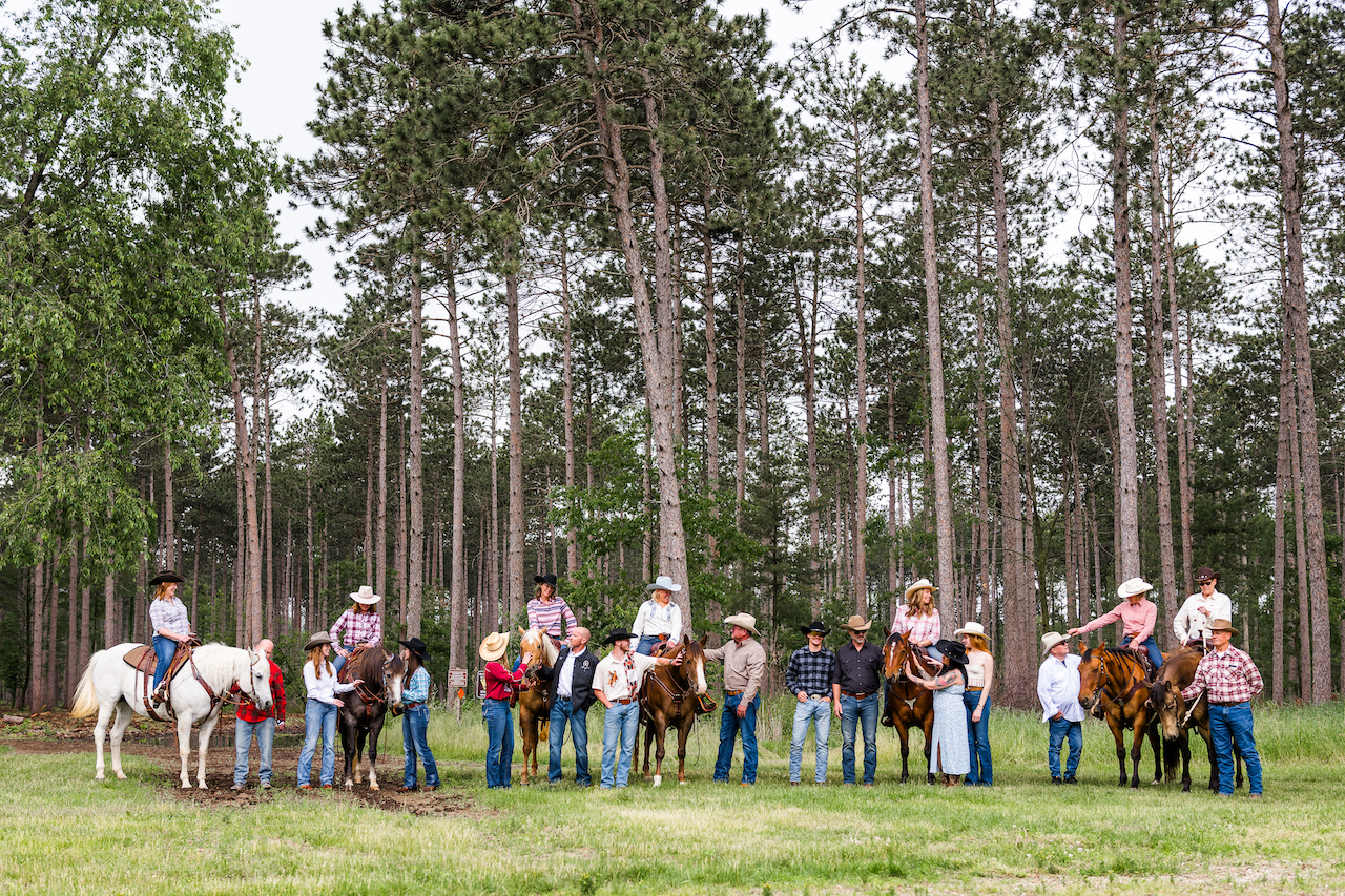 Multi-generational family with 20 people and 7 horses laughing and looking at each other surrounded by trees