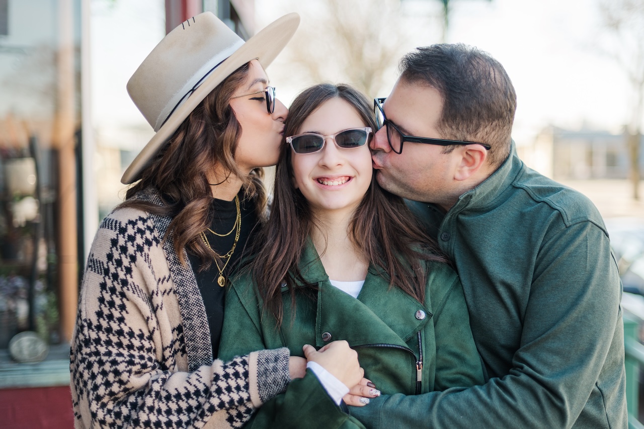 Alekx and Tom kissing their daughter Charlie on the cheek while wrapping their arms around her in a loving embrace.