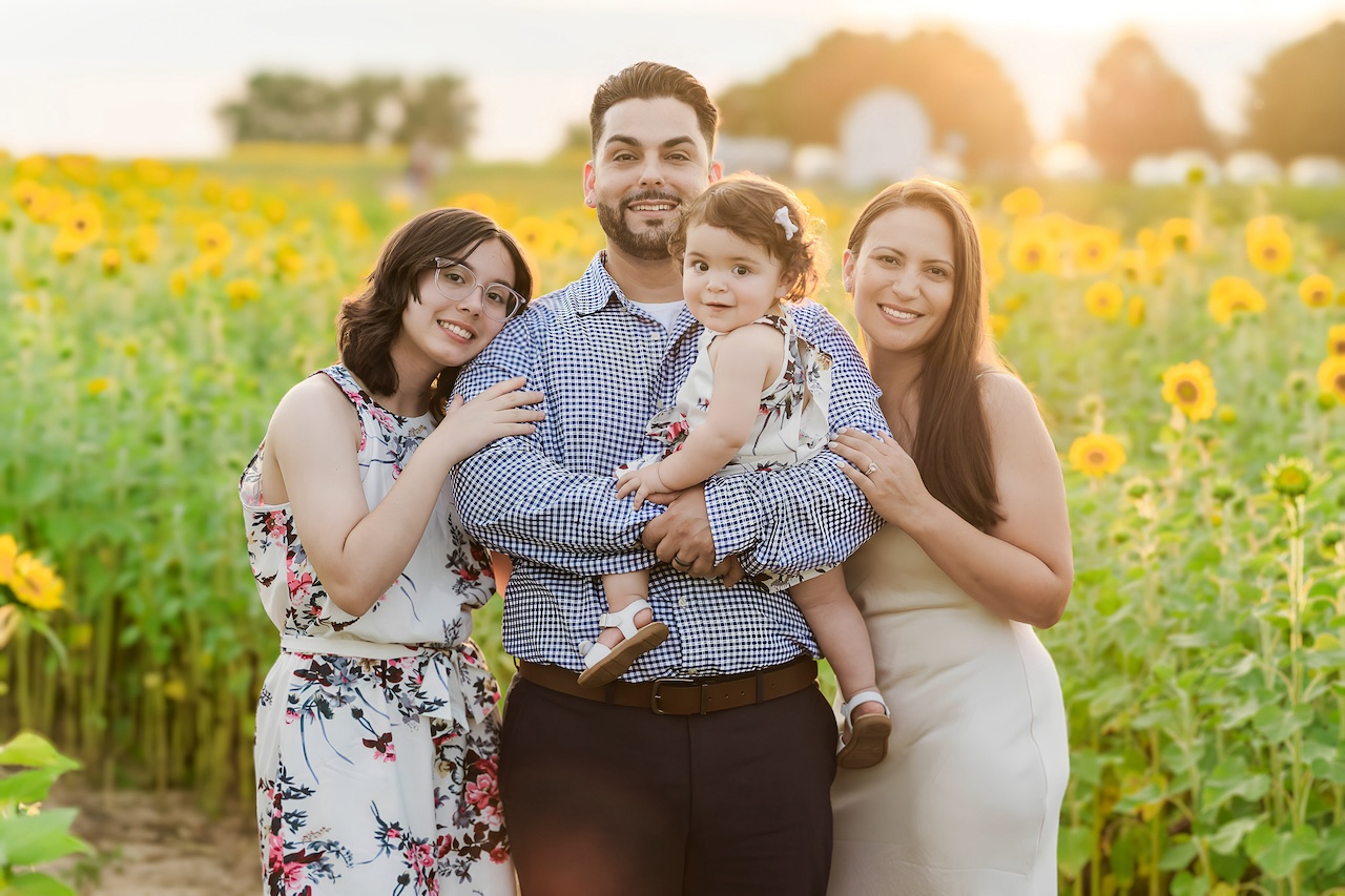 The Rodriguez family of four stands in a sunflower field at sunset, smiling with their arms around each other as the golden sunlight glows behind them.